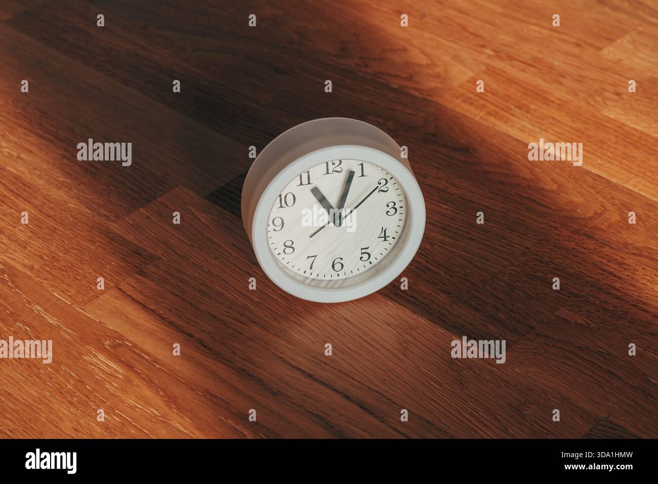 Une horloge analogique blanche simple repose seule sur des planches en bois chaudes, baignée par la lumière du soleil dorée dramatique avec une ombre longue et nette. Mise au point sélective. Banque D'Images