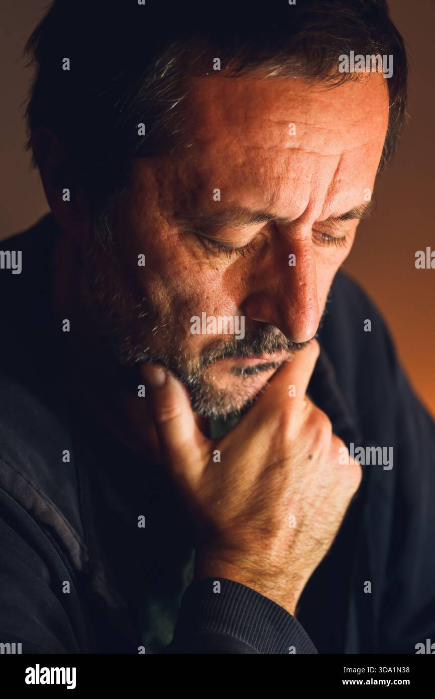 Homme inquiet assis seul dans une pièce sombre avec une lumière basse chaude, une expression sérieuse et une tension émotionnelle. Mise au point sélective. Banque D'Images