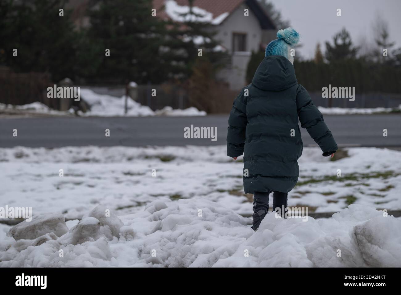 Un petit enfant vêtu de vêtements d'hiver marche à travers la neige fondante près d'une route de banlieue. La scène reflète la vie hivernale quotidienne, le temps froid, et vous Banque D'Images
