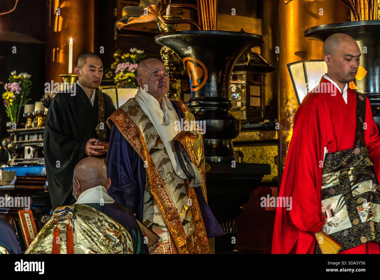 Cérémonie bouddhiste au temple Zenkoji à Nagano : un prêtre en chef, vêtu d'un magnifique kesa brodé d'or, tient une perle de prière juzu, flanquée de prêtres adjoints vêtus de noir et rouge. L'autel richement décoré en arrière-plan, avec des bougies, des offrandes florales et des objets rituels, est en harmonie avec le décor du Hondo, la salle principale de Zenkoji. Nagano, Préfecture de Nagano, Japon Banque D'Images
