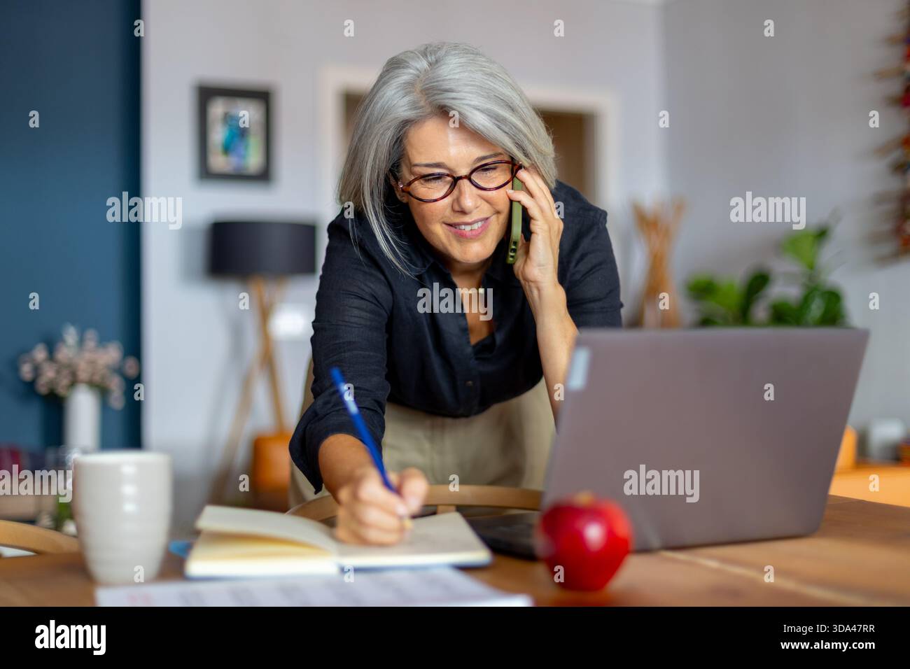 Femme d'affaires mature souriante s'engageant dans une conversation téléphonique tout en notant des notes et en travaillant efficacement à partir de son bureau à domicile Banque D'Images