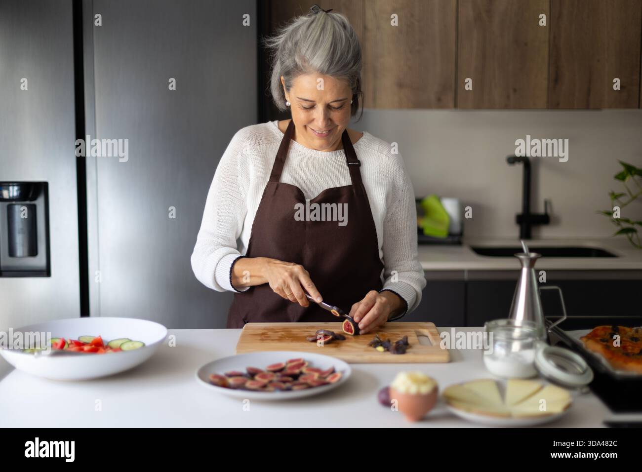 Heureuse femme mature portant un tablier préparant des aliments sains, coupant des figues sur une planche à découper, appréciant la cuisine à la maison Banque D'Images