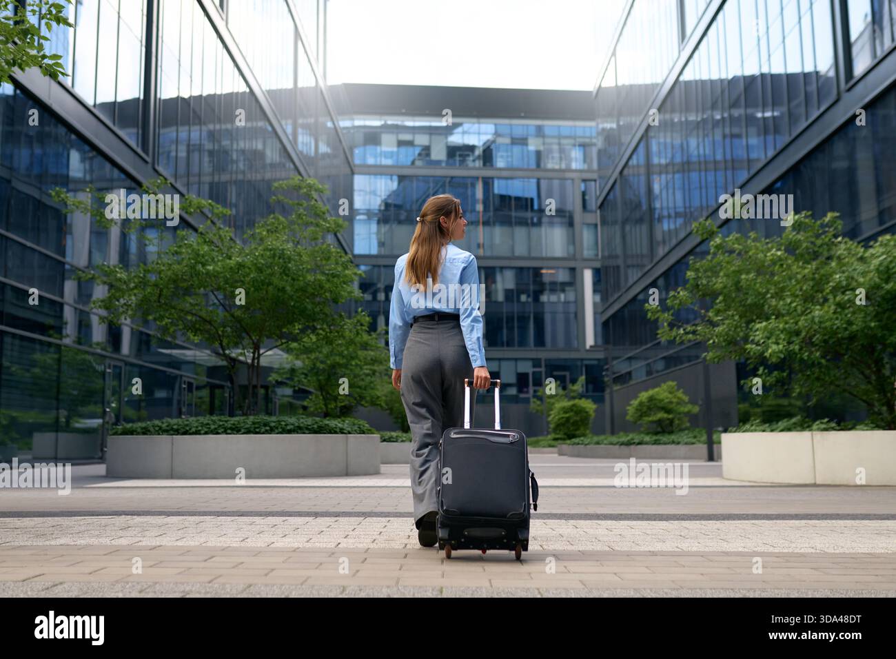 Vue arrière d'une femme confiante tirant une valise à travers un quartier d'affaires moderne. Bâtiments en verre réfléchissant, cadre urbain, concept de voyage, CA Banque D'Images