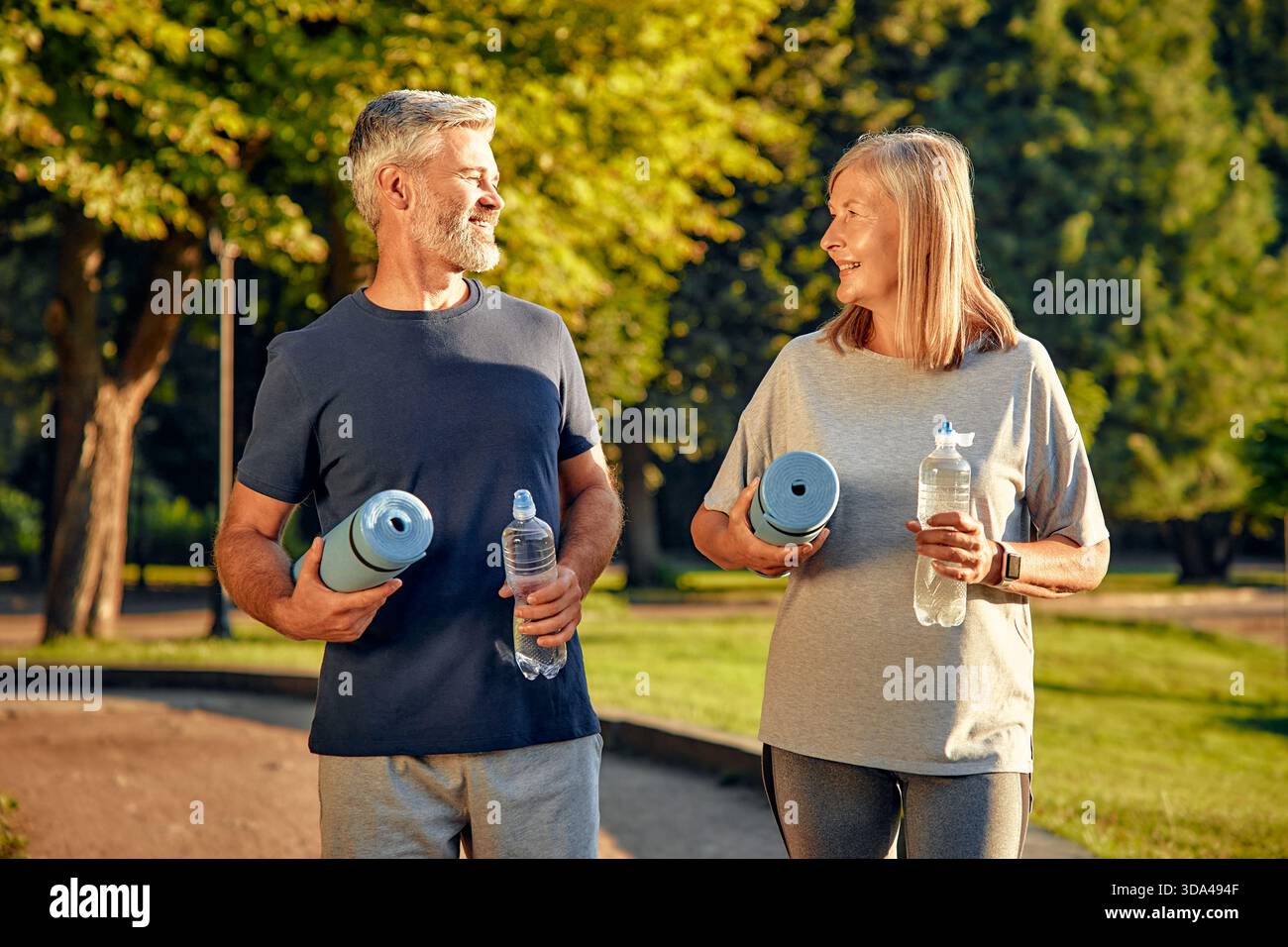 Un couple mature en forme et heureux profite d'une belle journée ensoleillée en plein air dans un joli parc, tenant joyeusement leurs tapis de yoga et bouteilles d'eau pendant eng Banque D'Images