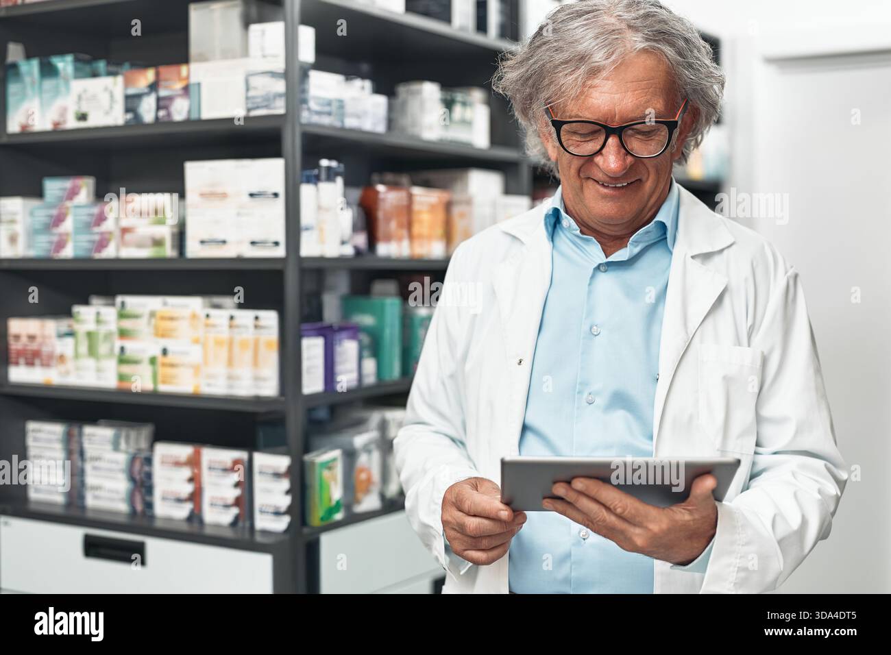 Pharmacien masculin âgé en manteau blanc et lunettes se tient dans une pharmacie, souriant tout en utilisant une tablette numérique contre les étagères avec la médecine et la santé Banque D'Images