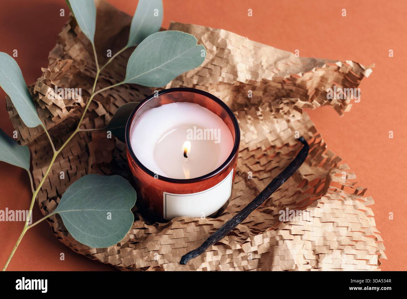 Bougie allumée en verre rouge avec des feuilles d'eucalyptus et du papier texturé sur fond orange chaud. Vue de dessus. Banque D'Images