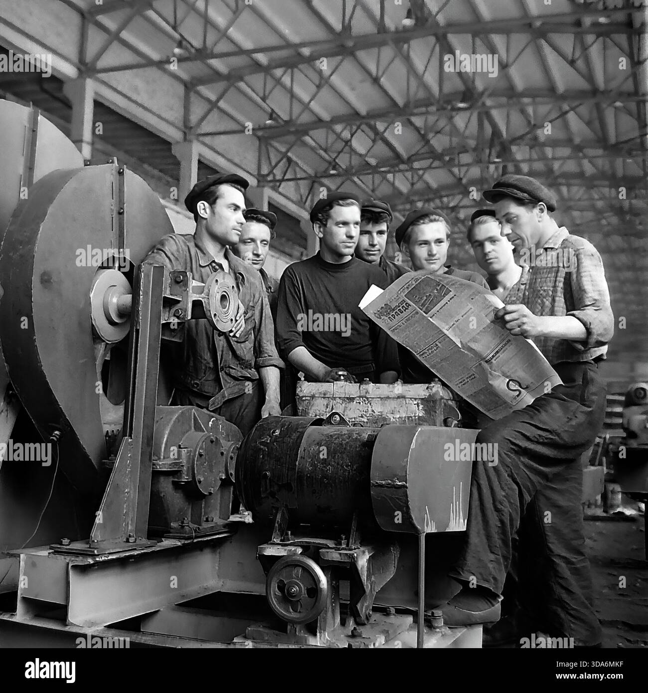 Slavyansk, RSS d'Ukraine, URSS - années 1970 : une brigade de travailleurs soviétiques dans l'atelier d'assemblage de l'usine de Stroymash lisant le journal Pravda pendant une pause. Les hommes en vêtements de travail et bérets sont rassemblés autour d'un grand ventilateur centrifuge industriel, discutant des dernières nouvelles. Cette photo d'archives en noir et blanc capture l'atmosphère de conscience politique et de vie collective de la paisible région du Donbass à l'époque soviétique. L'équipe de production de machines de l'industrie lourde prend un court repos. Banque D'Images