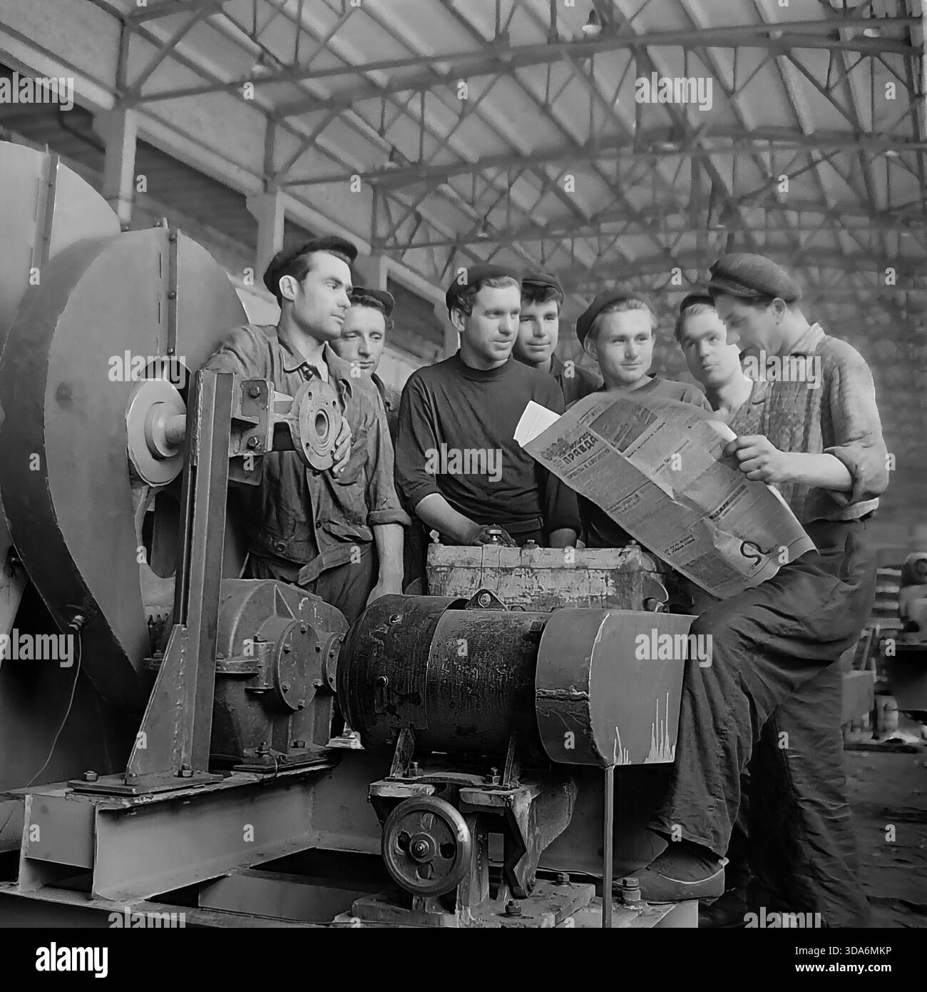 Slavyansk, RSS d'Ukraine, URSS - années 1970 : une brigade de travailleurs soviétiques dans l'atelier d'assemblage de l'usine de Stroymash lisant le journal Pravda pendant une pause. Les hommes en vêtements de travail et bérets sont rassemblés autour d'un grand ventilateur centrifuge industriel, discutant des dernières nouvelles. Cette photo d'archives en noir et blanc capture l'atmosphère de conscience politique et de vie collective de la paisible région du Donbass à l'époque soviétique. L'équipe de production de machines de l'industrie lourde prend un court repos. Banque D'Images