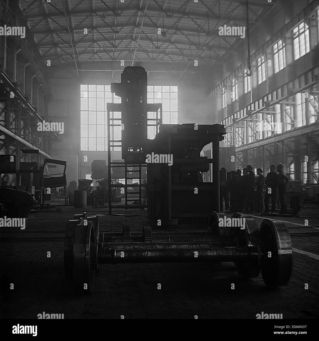 Slavyansk, RSS d'Ukraine, URSS - années 1970 : vue intérieure atmosphérique de l'atelier de l'usine de Stroymash avec une unité industrielle mobile massive en construction. De lourdes roues de chemin de fer sur un essieu se trouvent au premier plan, tandis qu'un groupe de travailleurs se silhouette au loin contre de grandes fenêtres. Cette photo d'archives en noir et blanc capture l'échelle colossale de la production de machinerie lourde et la puissance industrielle de la paisible région du Donbass à l'époque soviétique. Site de fabrication. Banque D'Images