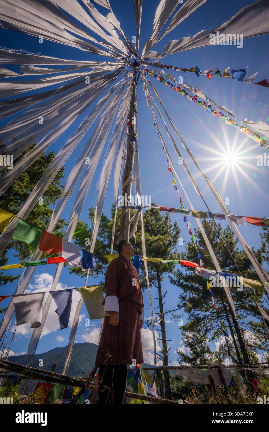Vue de drapeaux de prière flottant autour d'un haut poteau sous un soleil éclatant, avec une personne en tenue traditionnelle observant d'en bas, col de Kikila, Bumthan Banque D'Images