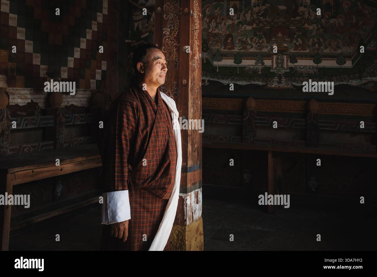 Vue d'un homme en vêtements traditionnels bhoutanais se dresse contre des murs peints de façon complexe, une étude dans la lumière et l'ombre, Trongsa, Trongsa, Bhoutan. Banque D'Images