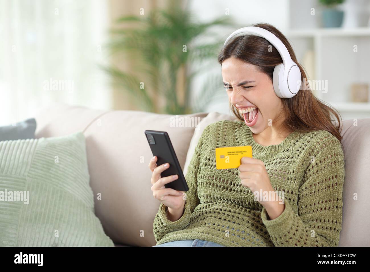 Femme excitée en vert portant casque achetant des médias en ligne avec carte de crédit et ordinateur portable assis sur un canapé à la maison Banque D'Images