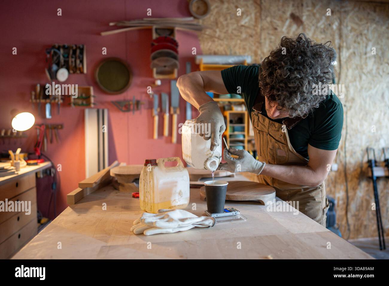 Menuisier homme préparant pour le travail sur le nouvel objet de mobilier, mesure précisément la quantité requise de résine époxy sur les balances électroniques à la table, versant du liquide dans la tasse en papier de la boîte, processus d'atelier. || autorisation du modèle Banque D'Images