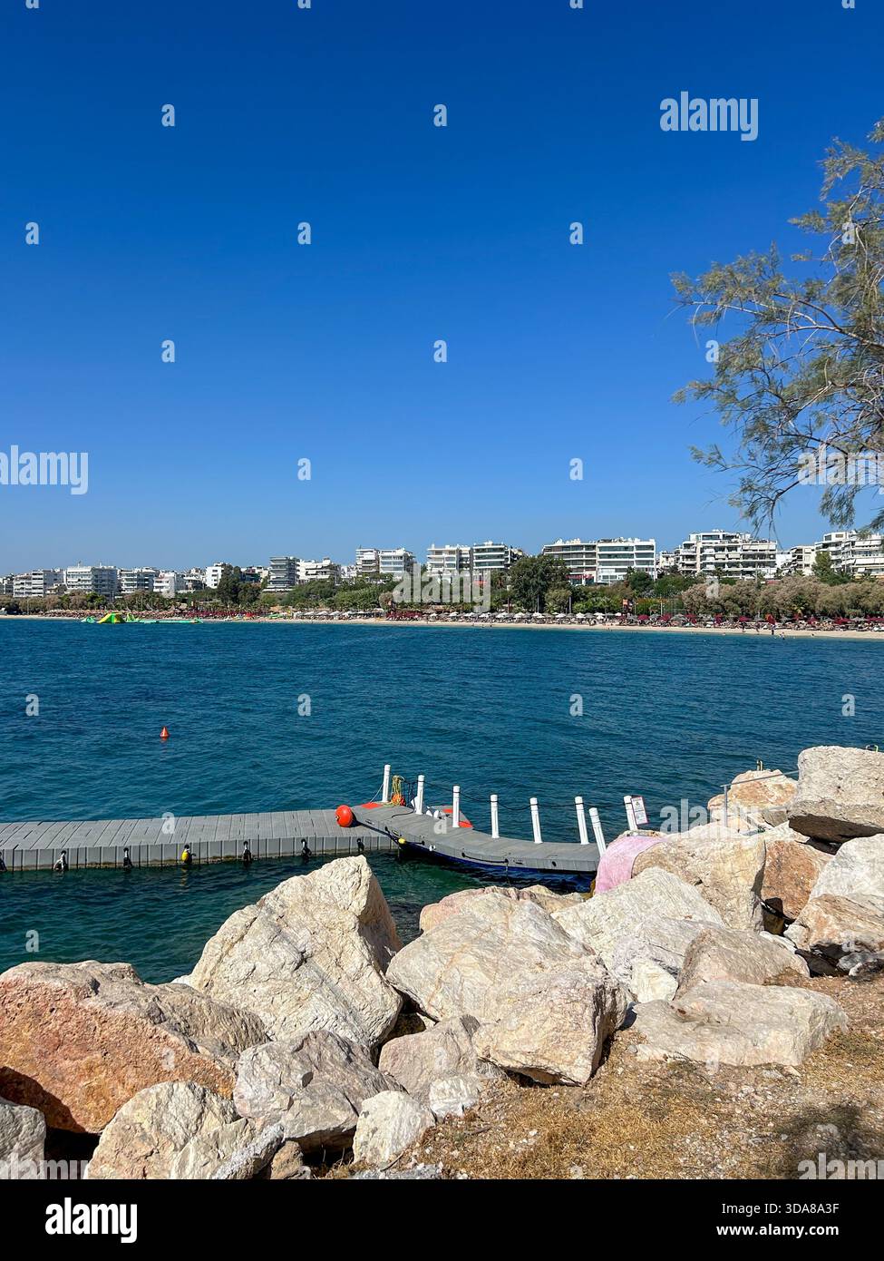 Vue côtière ensoleillée d'Alimos, un quartier balnéaire d'Athènes, avec des eaux bleues claires, un petit quai de marina, rivage rocheux et front de mer moderne. - Image de stock capturée avec un smartphone