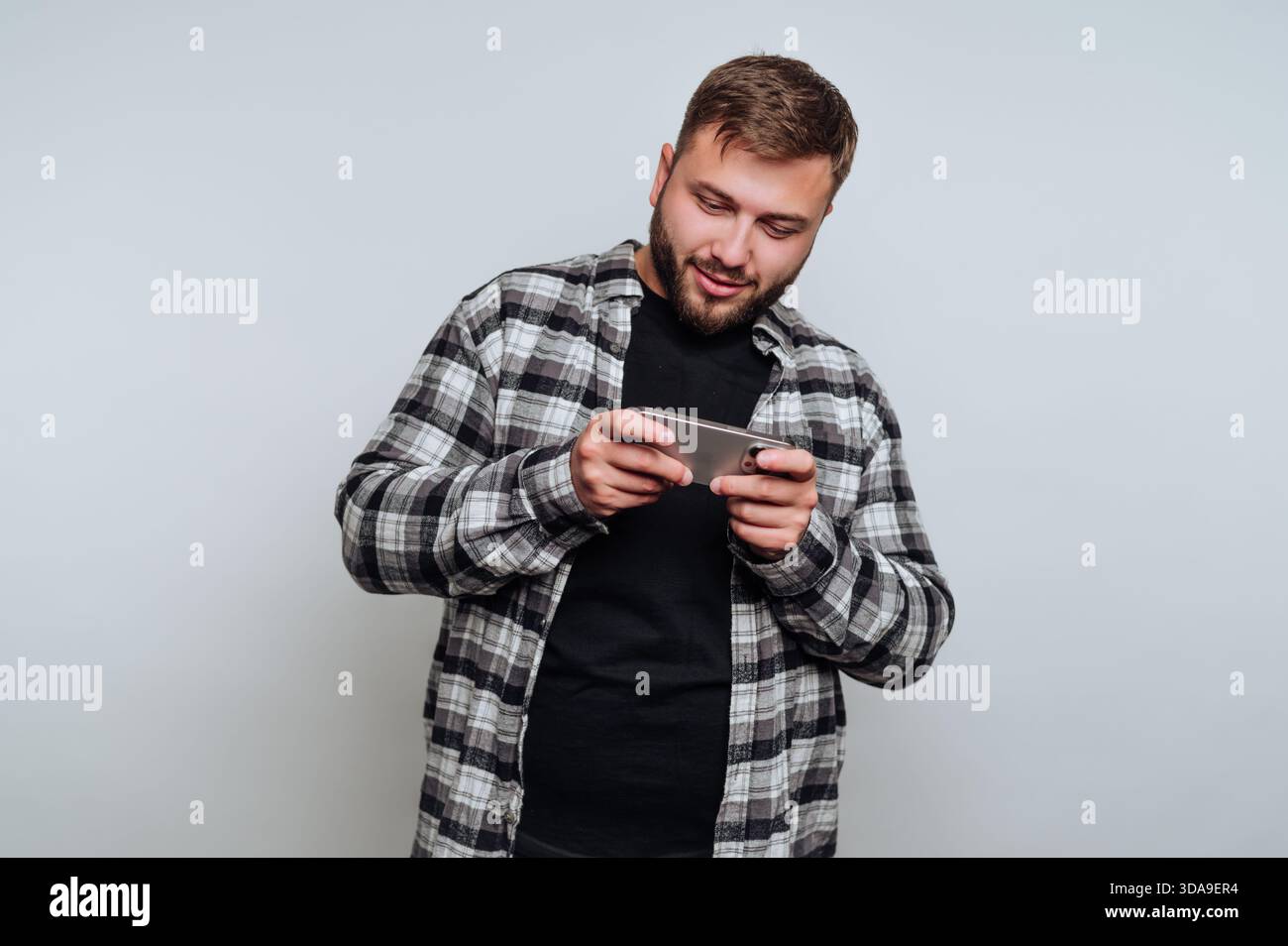 Un jeune homme dans une chemise à carreaux se tient à l'intérieur, concentré sur son smartphone comme il aime jouer. Il semble détendu et engagé dans son activité, smilin Banque D'Images