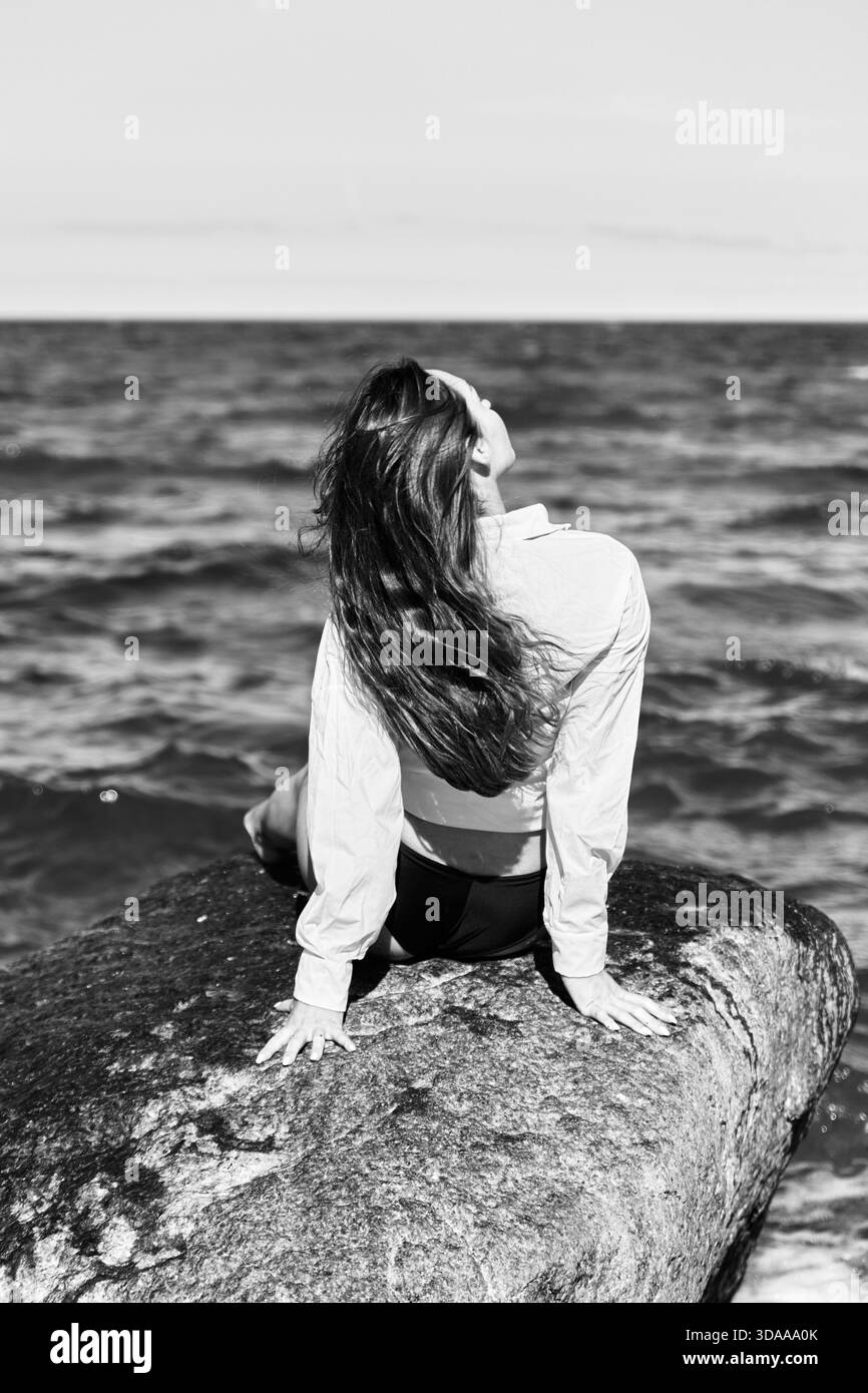 Jeune femme pose avec la tête inclinée en arrière sur un grand rocher près de la mer Baltique sous la lumière naturelle. Les tons noir et blanc soulignent l'ambiance sereine et le fond marin Banque D'Images