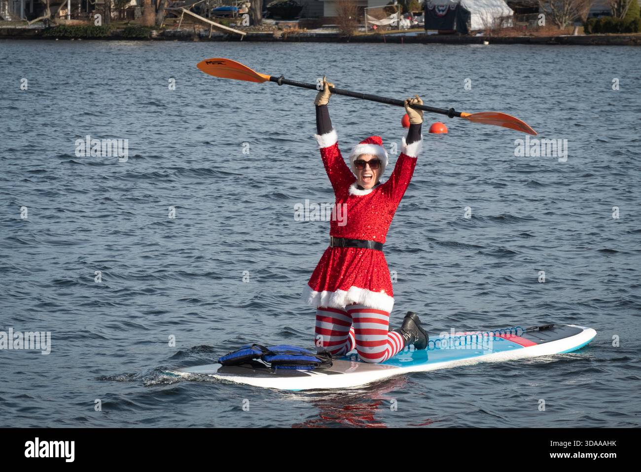 Lors de la célébration de Santa Palooza de 2025, une femme jubilante dans une tenue de Père Noël sur une planche de pagaie agite sa pagaie. À Greenwood Lake dans le comté d'Orange, NY. Banque D'Images