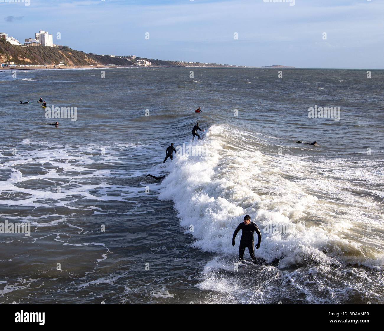 Plusieurs surfeurs chevauchent et attendent les vagues dans l'océan à Bournemouth UK avec un littoral lointain sous un ciel partiellement nuageux. Décembre surf hivernal Banque D'Images