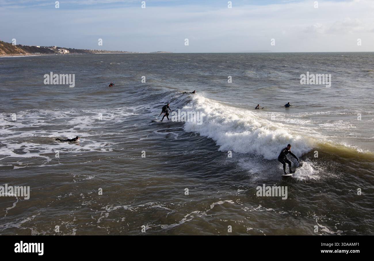 Plusieurs surfeurs chevauchent et attendent les vagues dans l'océan à Bournemouth UK avec un littoral lointain sous un ciel partiellement nuageux. Décembre surf hivernal Banque D'Images