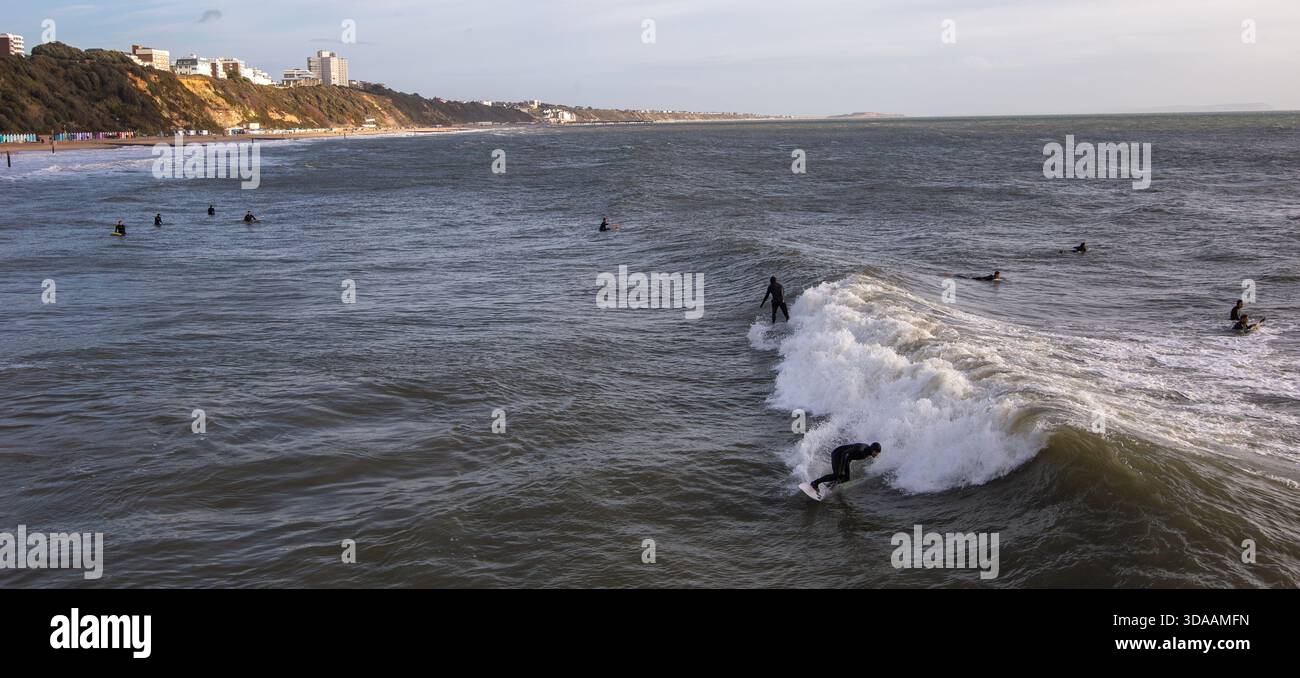 Surfeurs chevauchant des vagues près de la ville côtière de Bournemouth Royaume-Uni avec des falaises et des bâtiments visibles en arrière-plan sur une journée nuageuse de décembre Banque D'Images