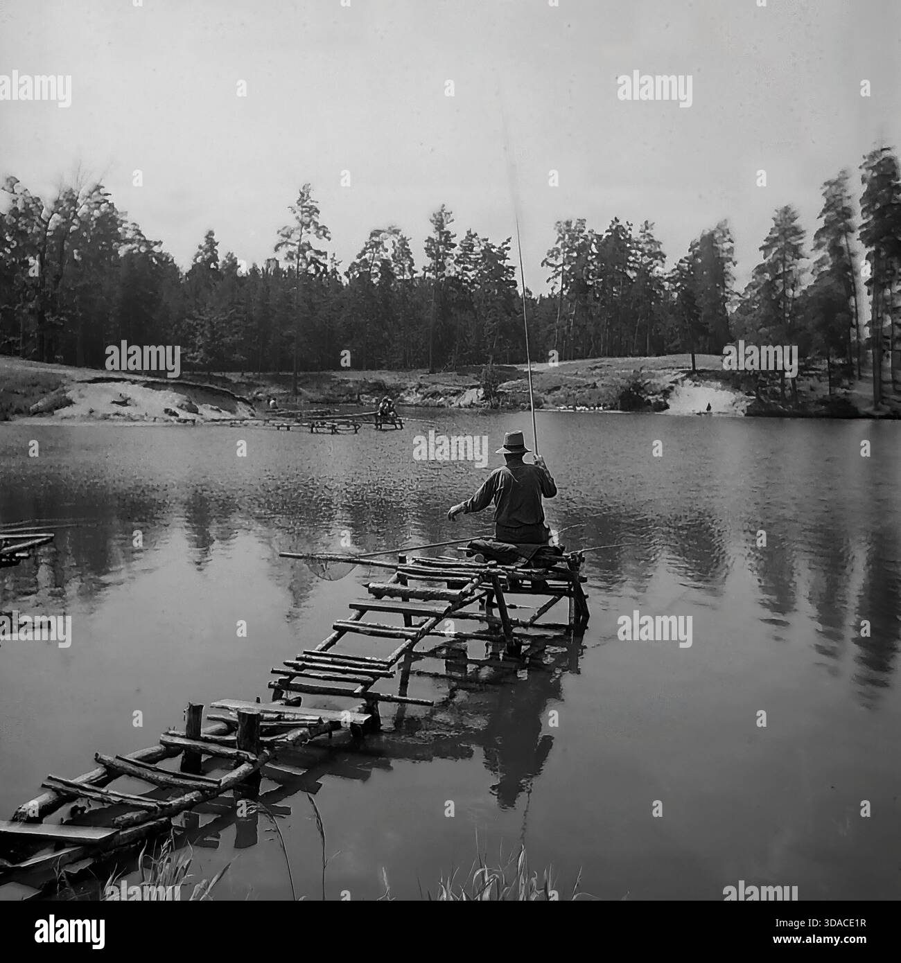 Une photographie pittoresque historique en noir et blanc des années 1960 capturant un pêcheur isolé sur le réservoir Red-Oskol (Krasnooskolskoye) près de Yatskoye, région de Donetsk, Ukraine. L'homme, portant un chapeau, est assis sur une jetée en bois rustique faite de bûches, pêchant avec une longue canne. L'arrière-plan présente une surface d'eau calme et une dense forêt de pins sur la rive opposée, reflétant la nature paisible et les traditions de loisirs du Donbass à l'époque soviétique. Banque D'Images