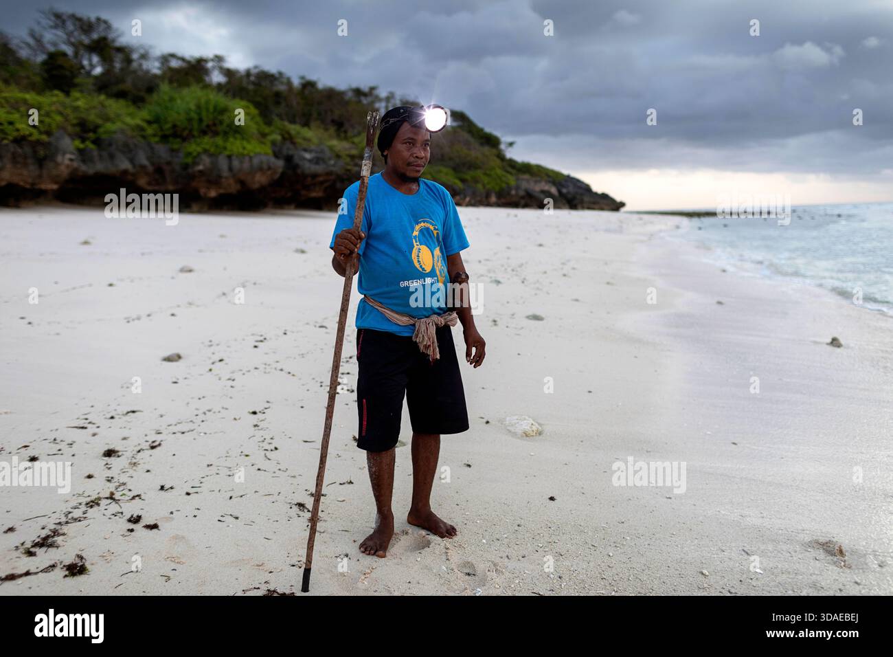 Un pêcheur local équipé d'une lampe frontale et d'une lance se préparant pour une expédition de pêche sur des récifs nocturnes sous un ciel orageux dramatique sur l'île de Sumba, en Indonésie. Banque D'Images