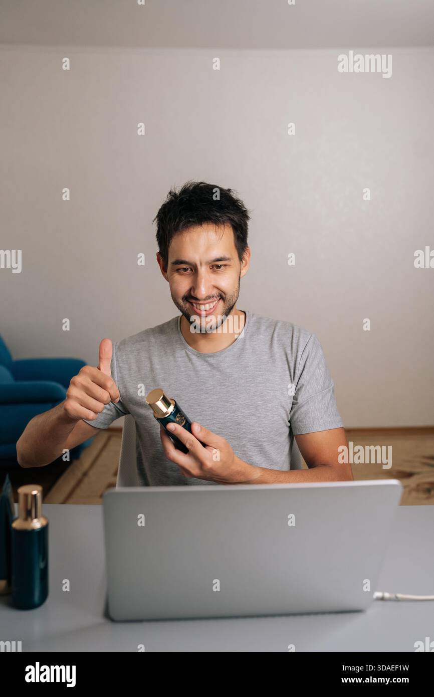 Vlogger masculin joyeux donnant les pouces vers le haut et tenant le produit cosmétique pendant un appel vidéo en ligne, examinant les articles de beauté pour le public des médias sociaux ou en direct Banque D'Images