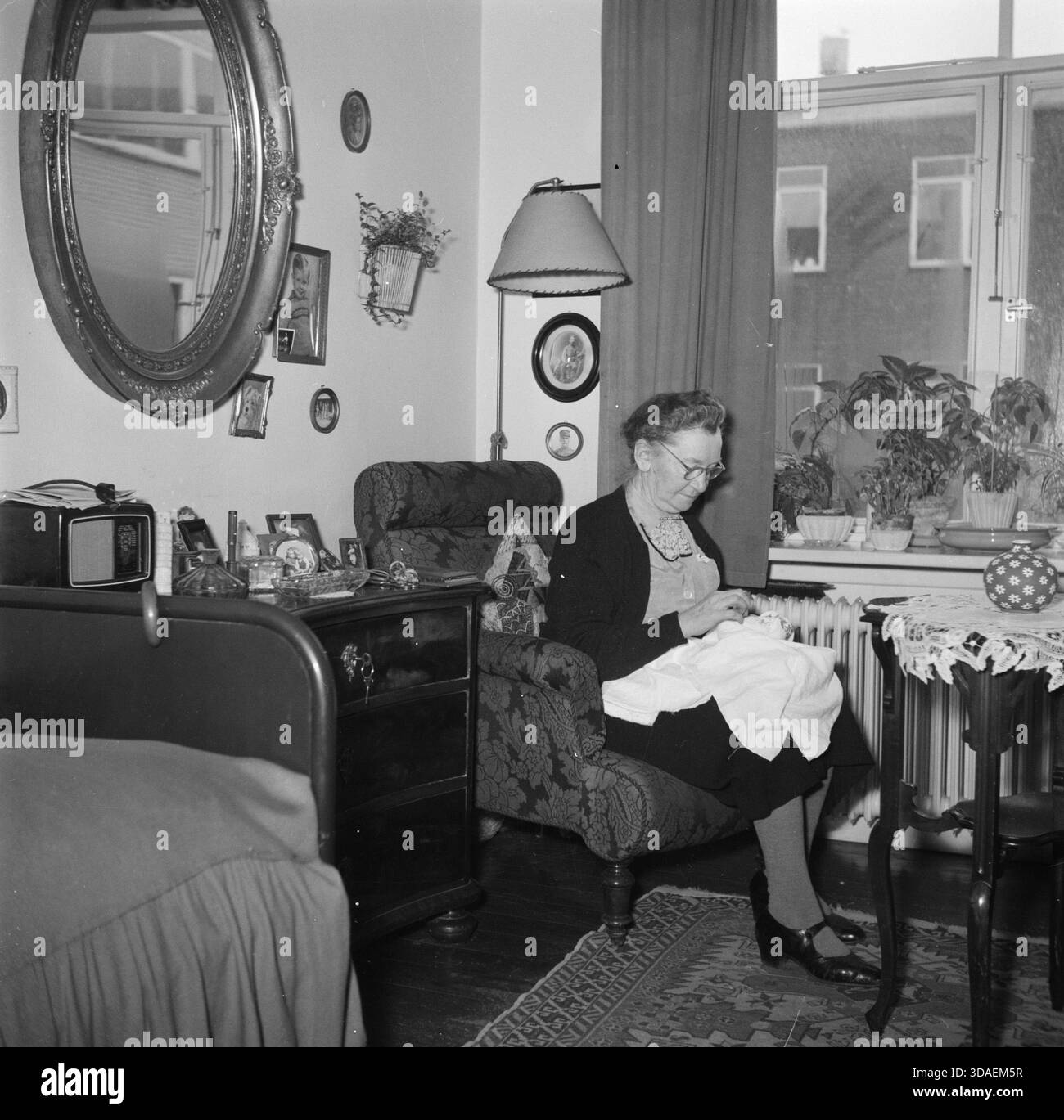 Femme âgée tricotant par la fenêtre (Danemark, mars 1954) : une femme âgée est assise sur une chaise près d'une fenêtre, travaillant tranquillement sur son artisanat, reflétant la vie quotidienne, les environnements de soins et le rôle des activités manuelles créatives pour les femmes dans les maisons de personnes âgées au milieu du XXe siècle au Danemark. Banque D'Images