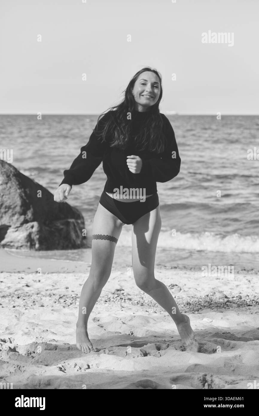 Jeune femme avec de longs cheveux foncés souriant franchement tout en se tenant pieds nus sur la côte sablonneuse de la mer Baltique. La lumière naturelle et la mer calme créent une atmosphère sereine Banque D'Images