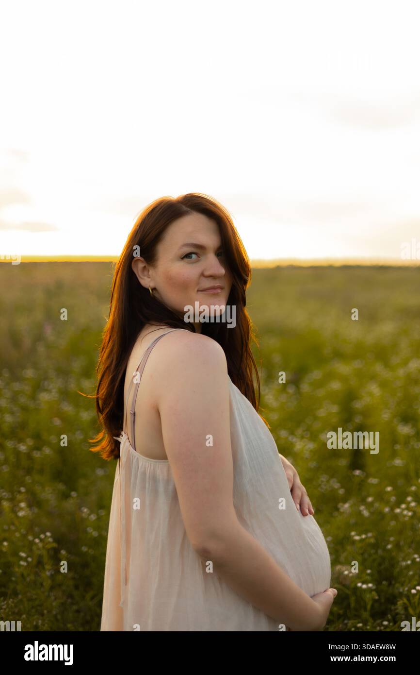 Une femme enceinte sereine se promène dans un champ de campagne ensoleillé, son regard rencontrant la caméra. Le moment paisible capture la beauté et l'anticipation Banque D'Images