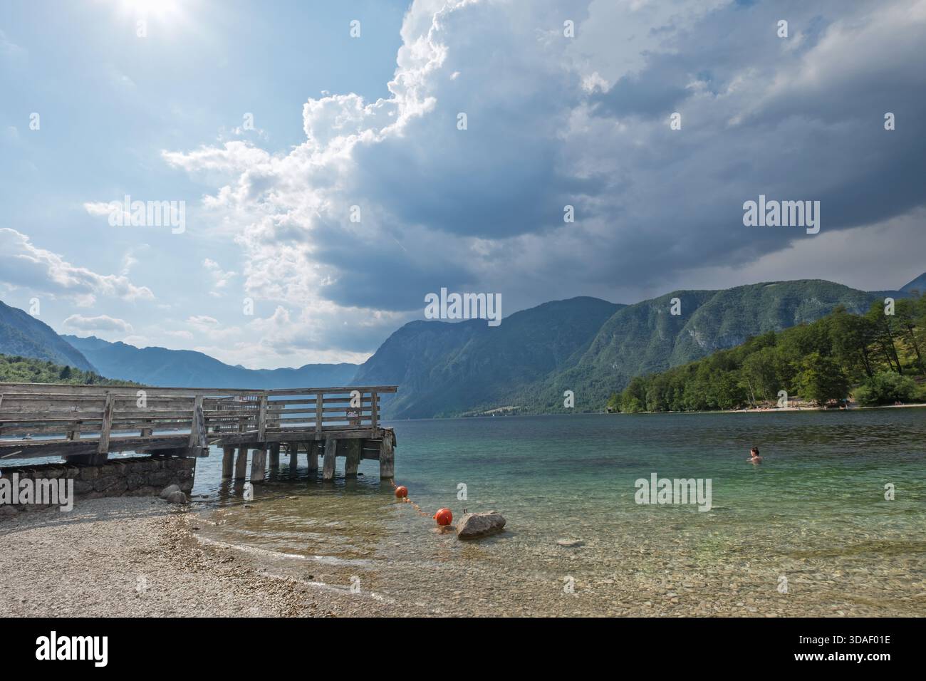 Lac turquoise avec jetée en bois, nageur, bouées oranges et ciel de montagne spectaculaire. Moment d'été alpin pittoresque avant la tempête. Banque D'Images