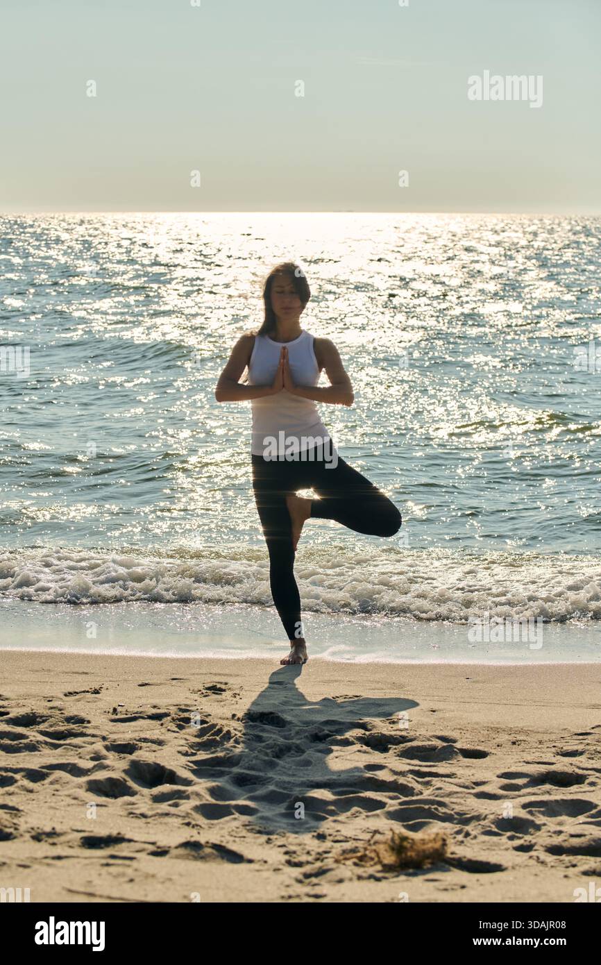 Une femme adulte effectue un exercice de yoga de pose d'arbre sur le rivage de sable de la mer Baltique avec de l'eau calme et la lumière du soleil chatoyante. Scène capture la pleine conscience et tranqui Banque D'Images