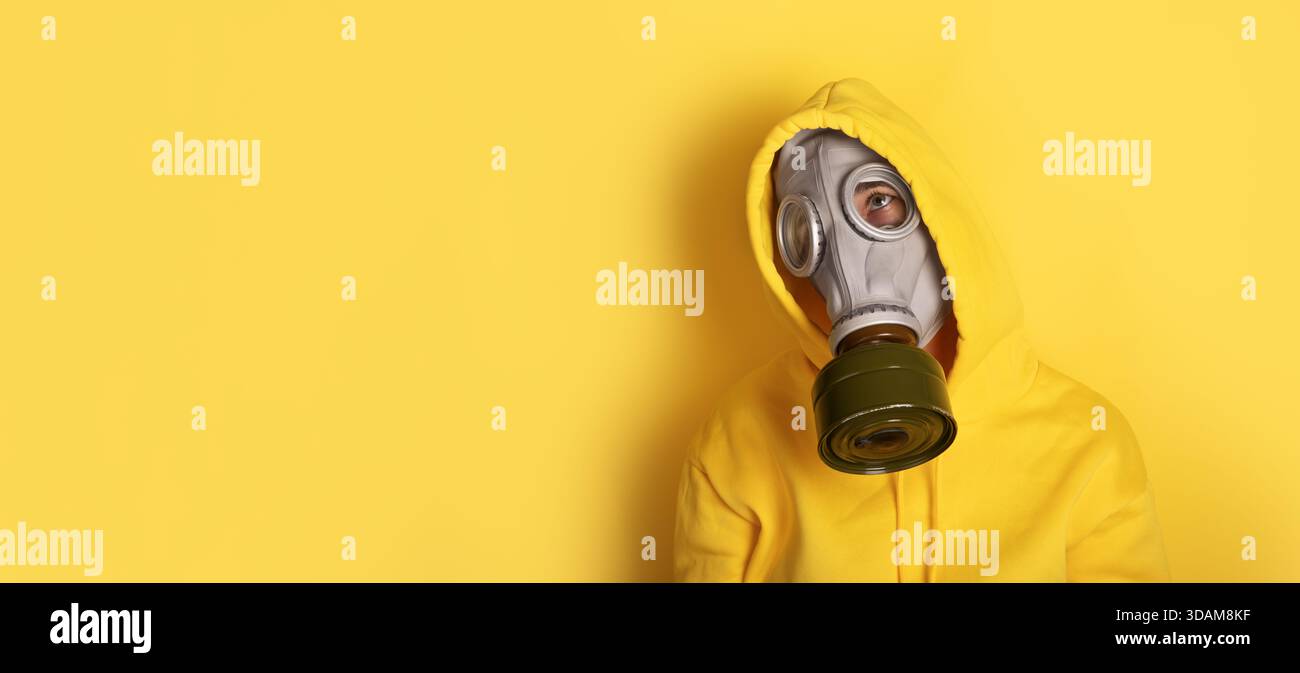 Individu portant un masque à gaz gris et un sweat à capuche jaune sur un fond jaune Uni, offrant un fort contraste visuel et un espace de copie. Banque D'Images