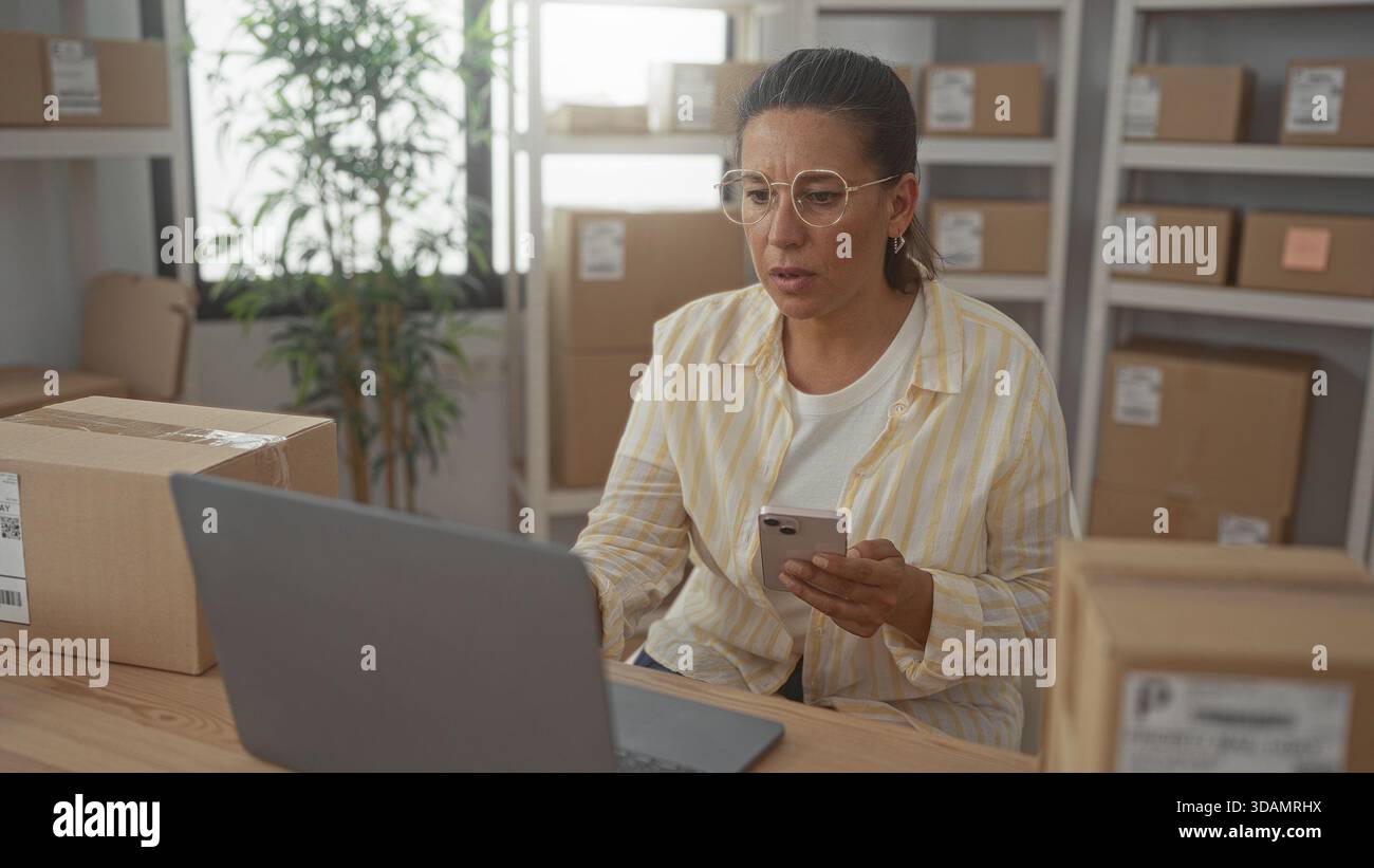 Femme tenant le téléphone avec la main droite, tapant sur ordinateur portable, entourée de boîtes d'expédition en carton dans le bâtiment ; concernent la logistique des petites entreprises. Banque D'Images