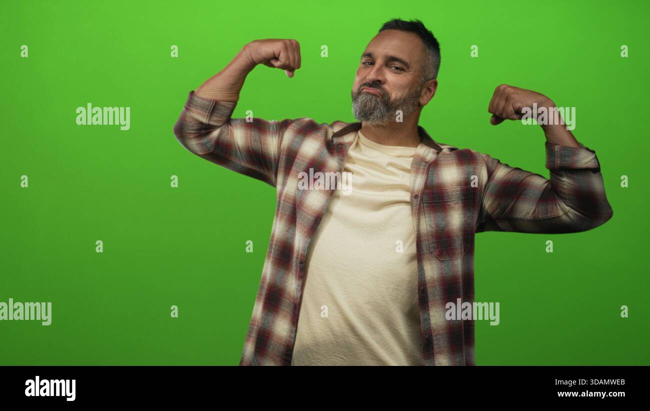 Homme hispanique d'âge moyen avec les cheveux gris et la barbe fléchit les bras et crie dans le studio vert ; énergie de force de confiance. Banque D'Images