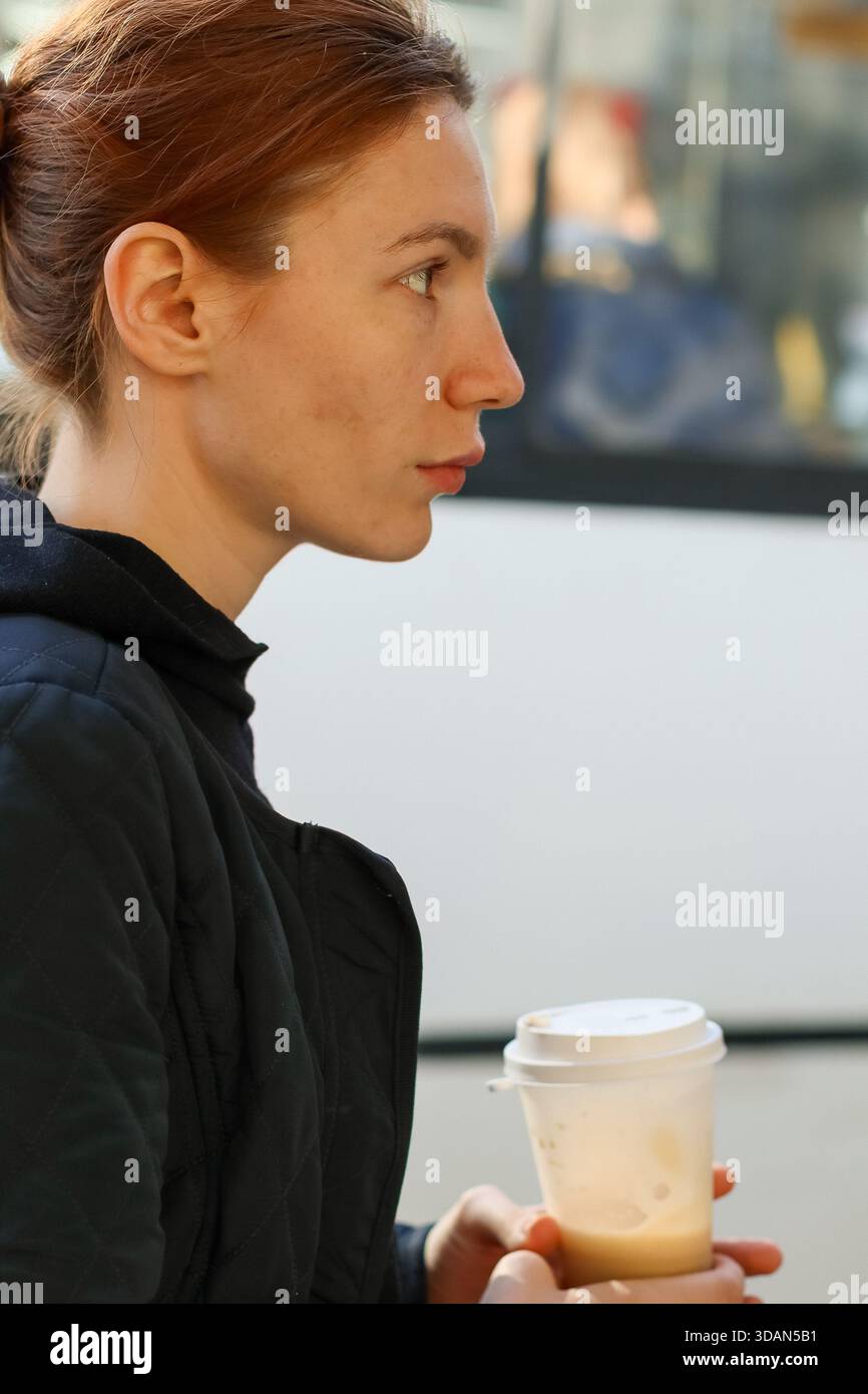 Gros plan d'une jeune femme réfléchie avec queue de cheval serrant le café à emporter près d'un bus public en milieu urbain Banque D'Images