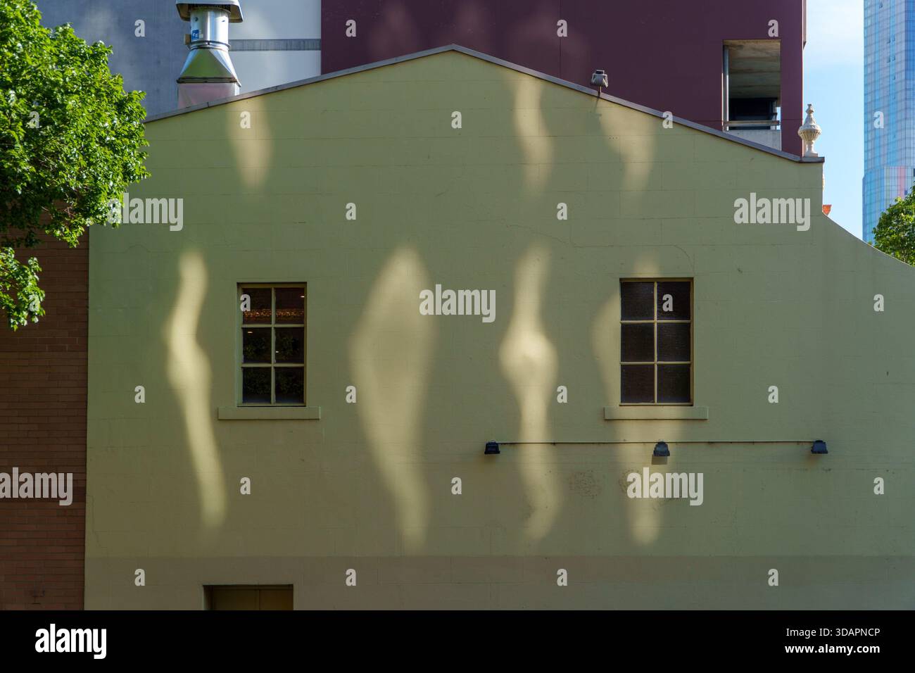 La lumière du soleil projette des ombres spectaculaires et ondulantes sur une façade de bâtiment vert pâle, avec deux fenêtres quadrillées, au milieu de structures urbaines variées. Banque D'Images