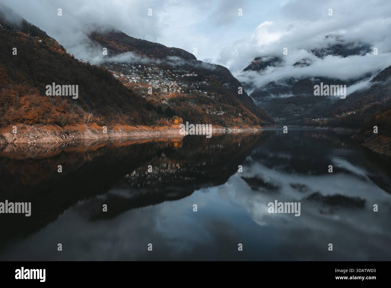 Vue sur un lac serein reflétant les spectaculaires montagnes voilées de nuages et les arbres d'automne colorés accrochés aux pentes, Verzasca, Tessin, Suisse. Banque D'Images
