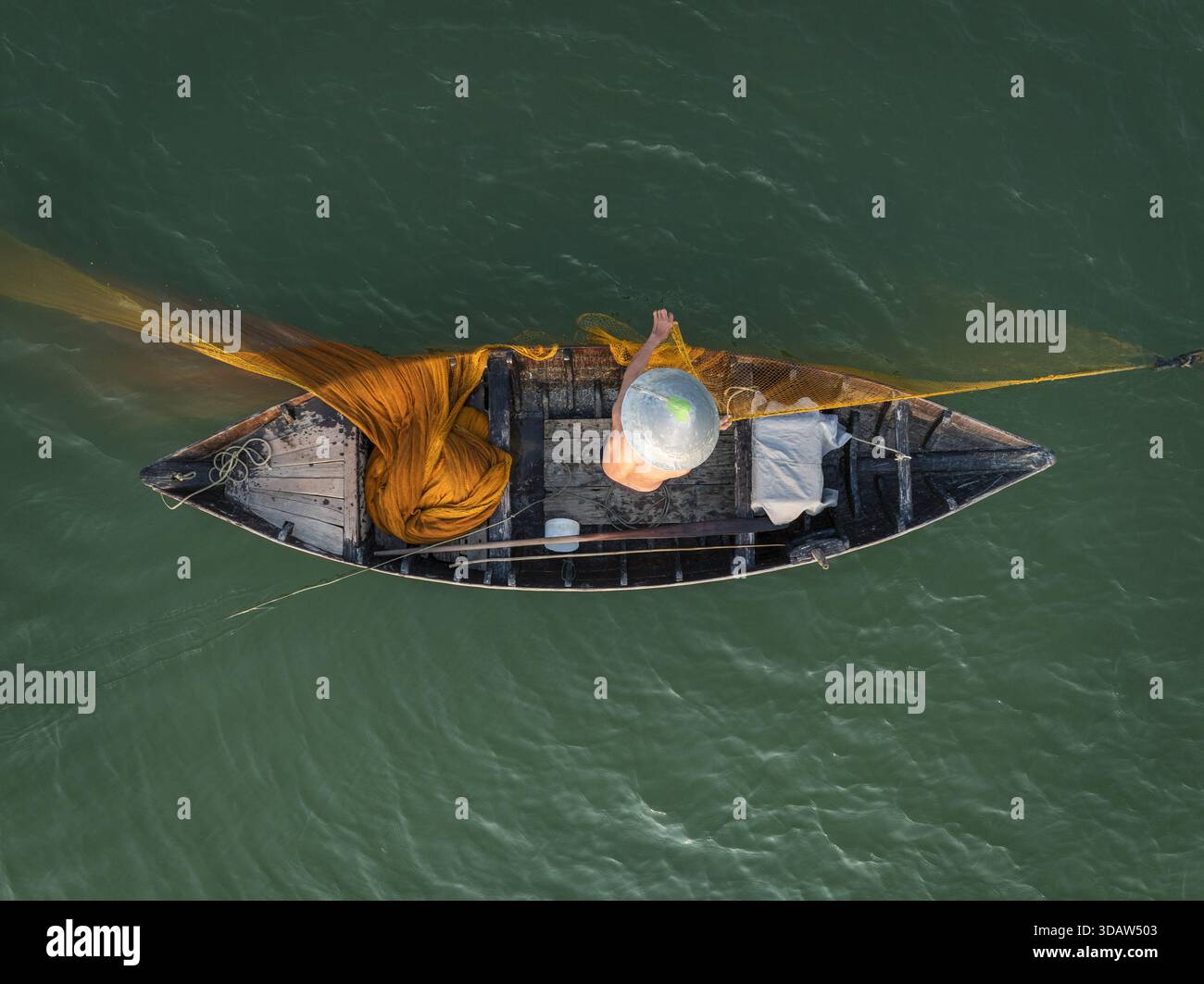 Vue aérienne d'un pêcheur solitaire jetant un filet jaune vif depuis son bateau altéré sur les eaux vertes ondulantes, Hội an, Quảng Nam, Vietnam. Banque D'Images