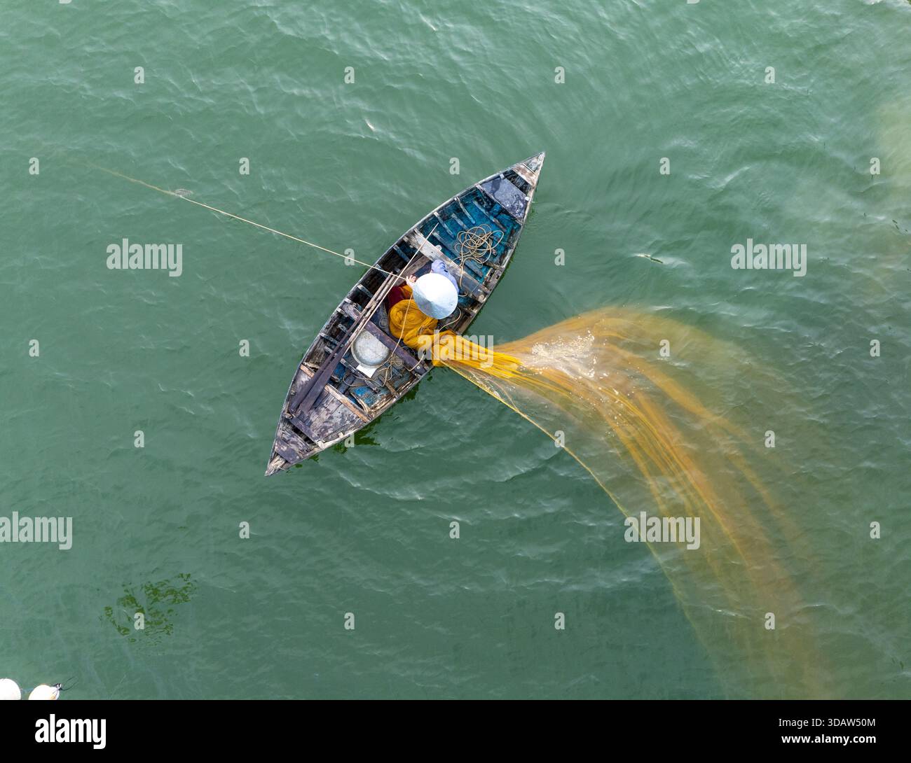 Vue aérienne d'un pêcheur solitaire projette son filet jaune éclatant depuis un bateau altéré, créant un contraste saisissant avec les eaux turquoises sereines, Banque D'Images