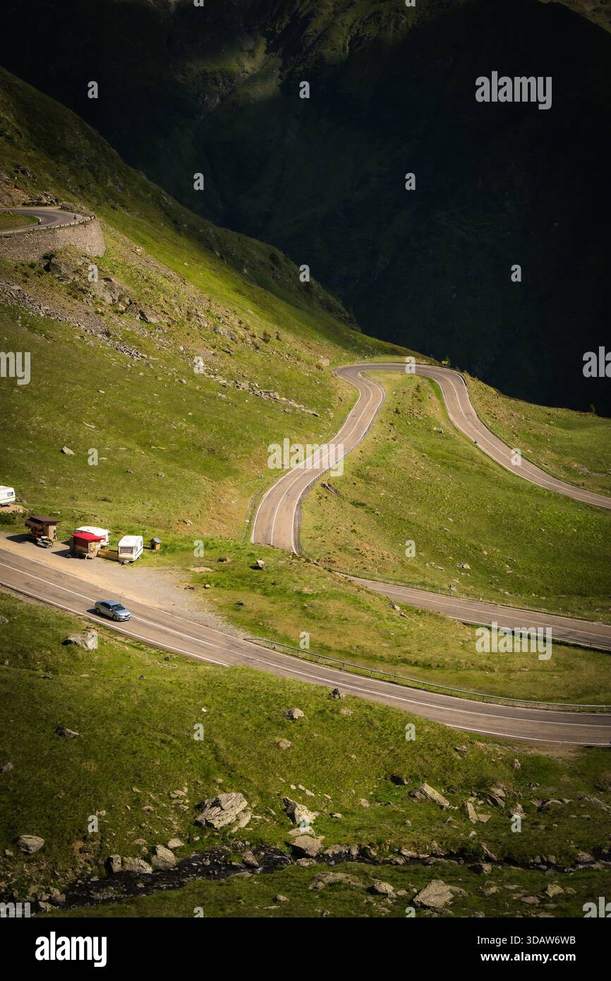 Vue d'une route sinueuse serpentant à travers des collines verdoyantes, voitures se déplaçant le long du chemin, illuminé par les rayons du soleil, Poienari, Transilvania, Roumanie. Banque D'Images
