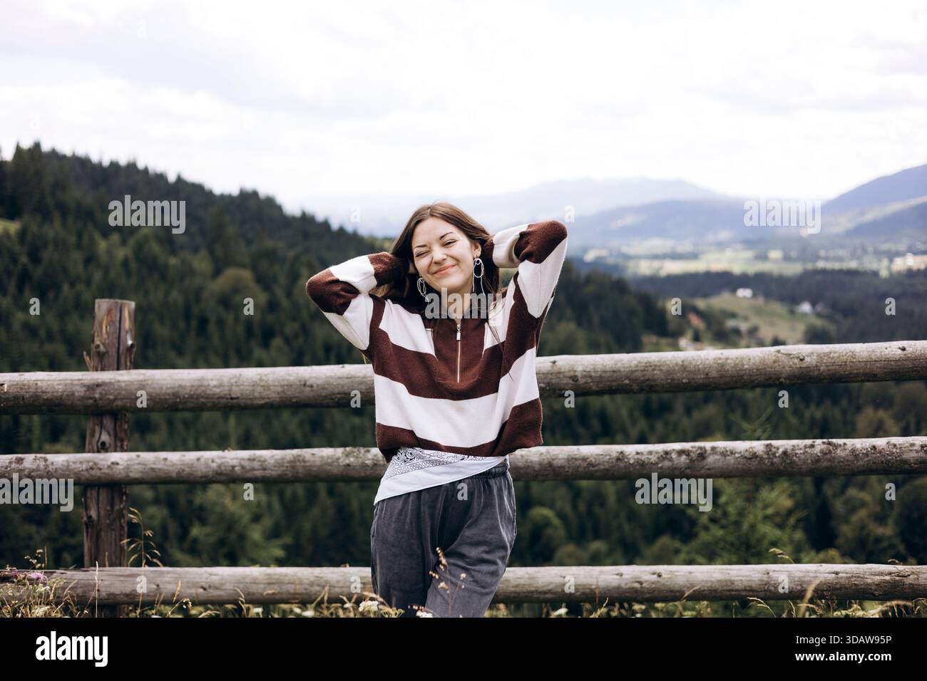 Jeune femme debout devant un paysage de montagne pittoresque, souriant joyeusement avec les mains derrière la tête. Une clôture en bois ajoute un charme rustique au s. Banque D'Images