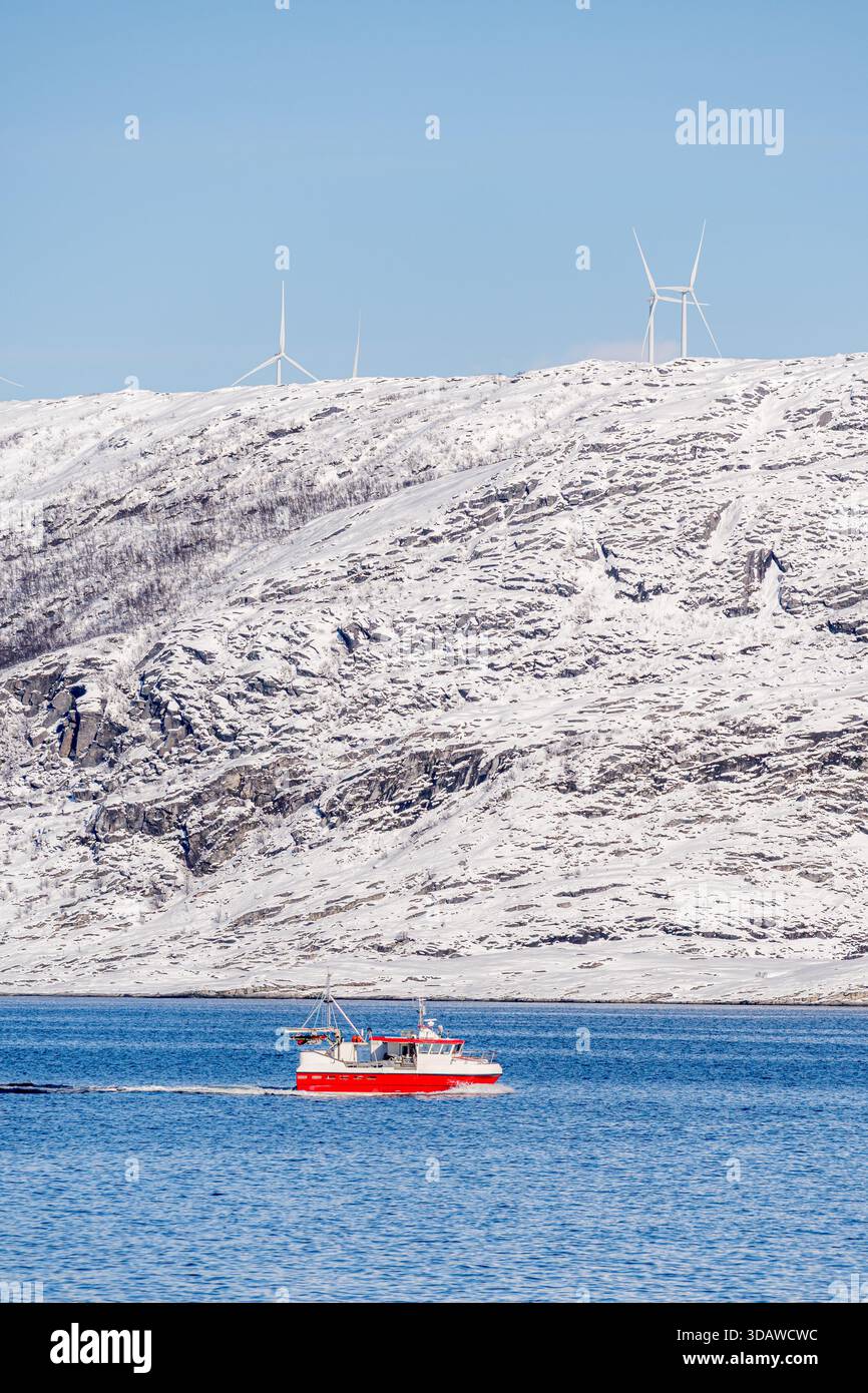 Un bateau de pêche rouge vif navigue dans les eaux bleues des fjords norvégiens, à Kvaloysletta, avec des collines enneigées et des éoliennes imposantes Banque D'Images