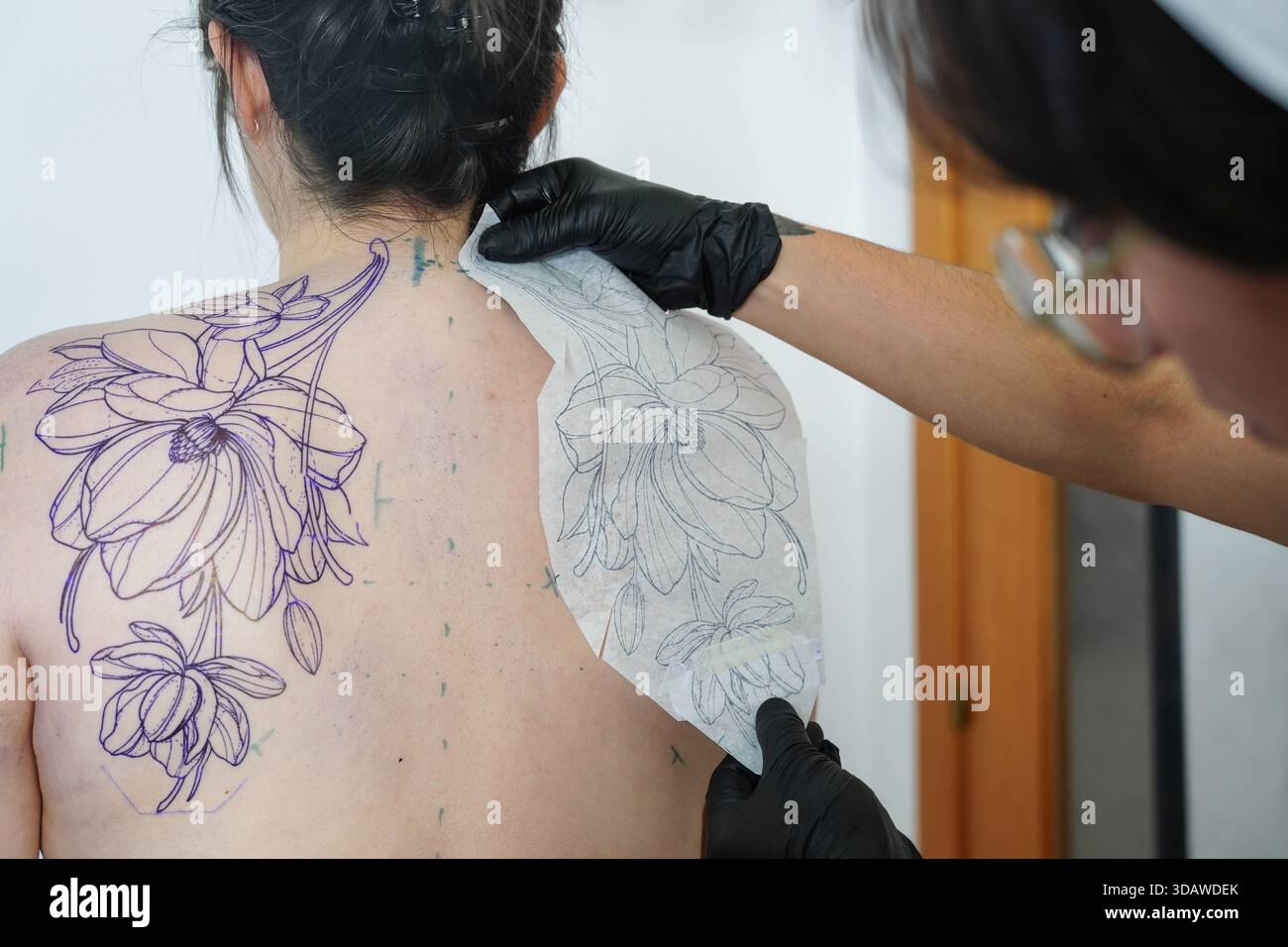 Un tatoueur féminin qualifié tatoue méticuleusement un dessin floral détaillé sur le dos d'un client. L'artiste utilise un pochoir pour assurer précision et acc Banque D'Images