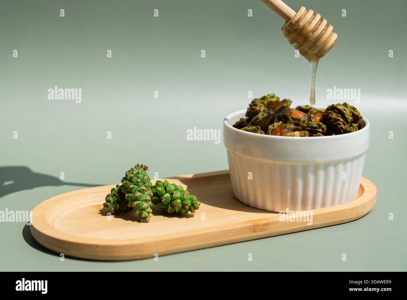 Le miel de pommes de pin bio coule d'une benne à miel en bois sur des pommes de pin vertes dans un bol en céramique blanche. Sur fond vert clair et bois Banque D'Images