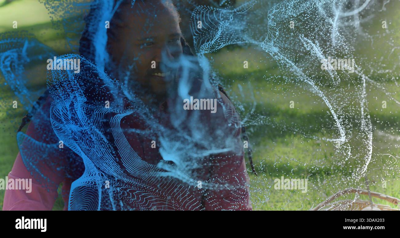 Homme assis avec des dreads en veste rouge souriant sur l'herbe du parc, hologramme bleu, panier et enfant Banque D'Images