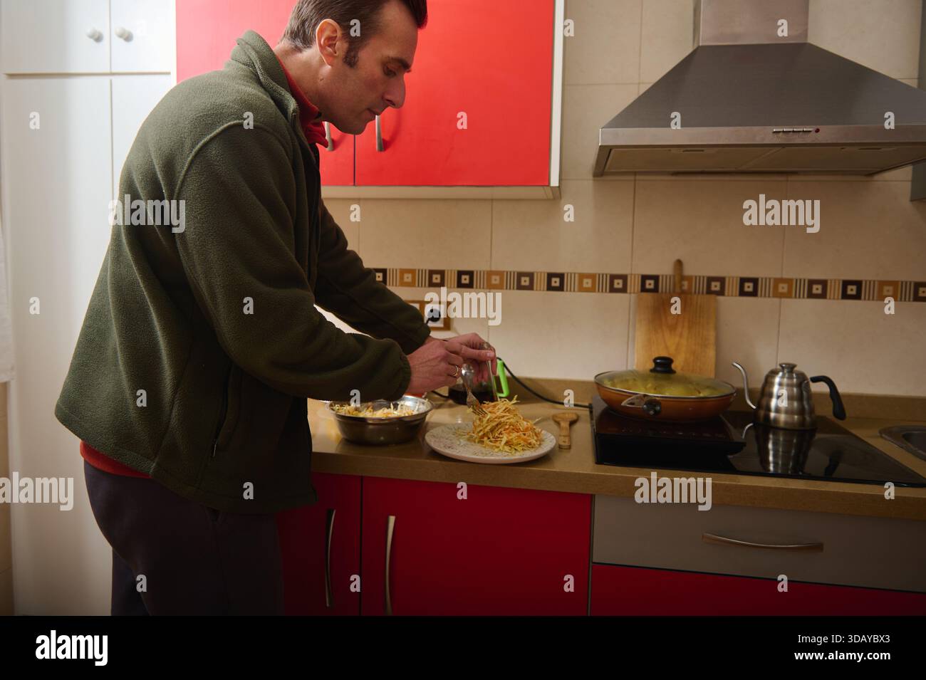 Un homme en veste verte prépare un repas dans une cuisine contemporaine. Il saupoudre du fromage sur des pâtes sur une assiette tandis qu'une casserole mijote sur le poêle. Banque D'Images