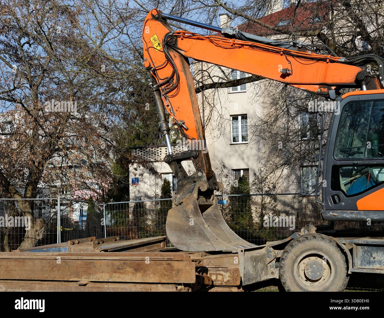 Vue latérale rapprochée du bras de pelle orange, des tuyaux hydrauliques et du seau métallique à côté de la clôture temporaire, fond de bâtiment résidentiel, lumière du jour brillante. Banque D'Images