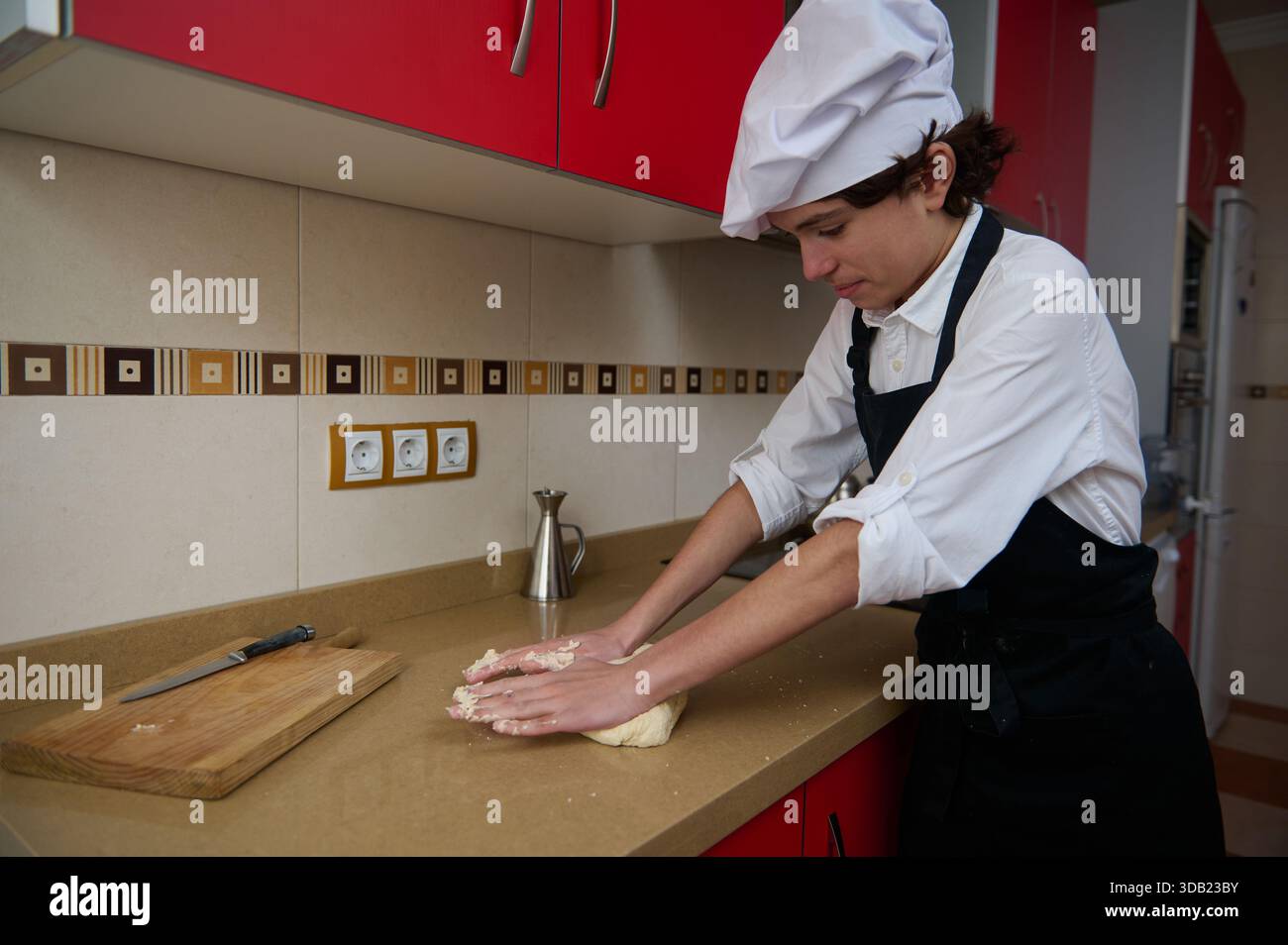 Un chef concentré en chemise blanche et tablier noir pétrit la pâte sur un comptoir beige, avec une planche à découper et un couteau à proximité dans une cuisine rouge vif. Banque D'Images
