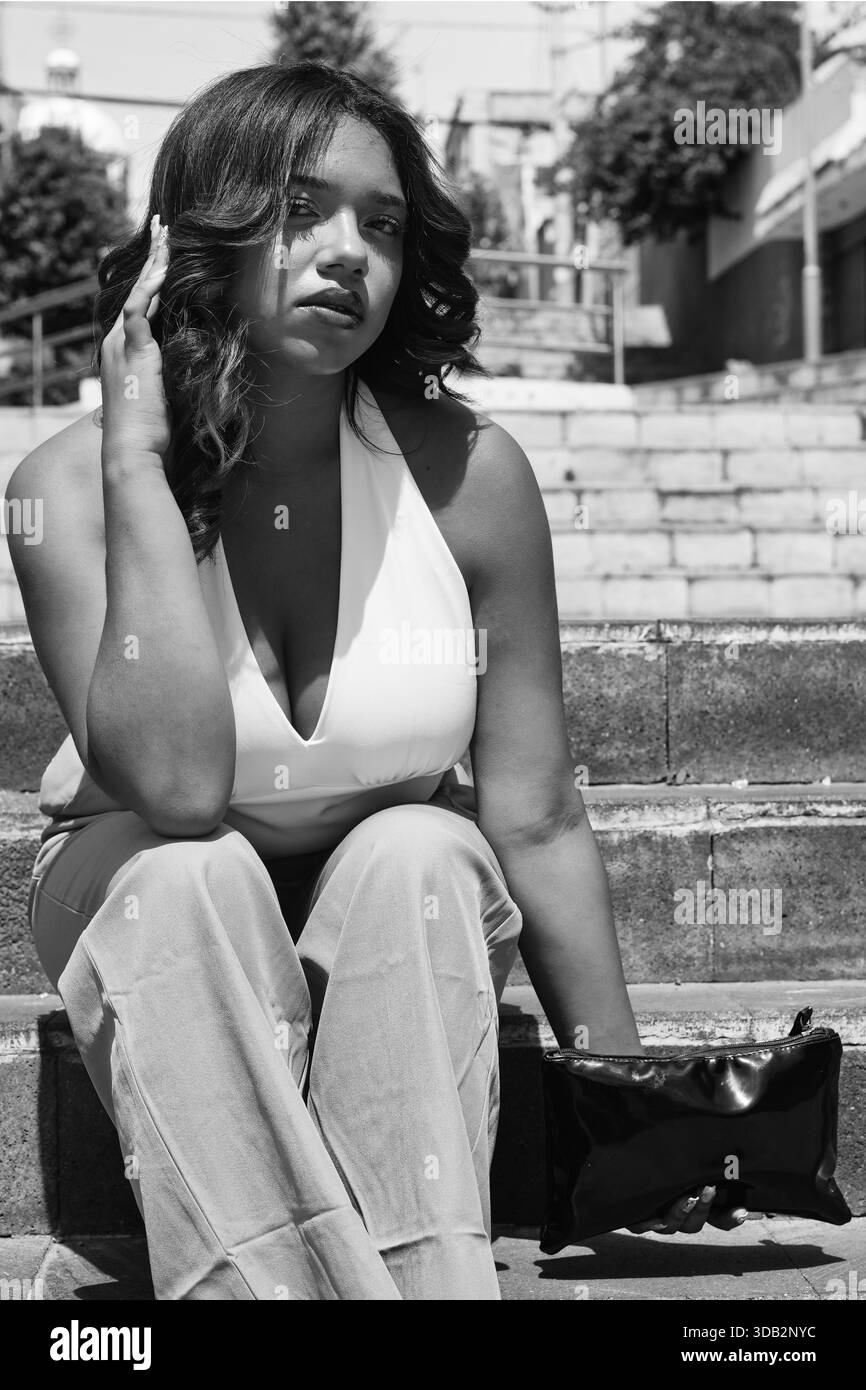Femme assise sur de vieux escaliers en béton, tenant une pochette noire et regardant la caméra avec une expression confiante, montrant le style personnel et urb Banque D'Images